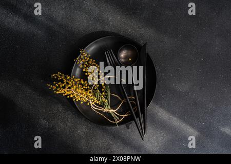 Top view of modern black table setting complete with plate, cutlery, and a bright yellow mimosa arrangement on a dark surface. Stock Photo