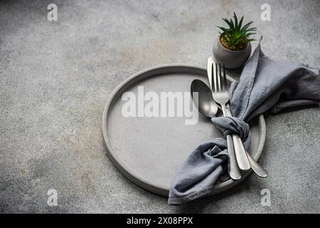 Top view of elegant, minimalist table setting featuring a simple ceramic plate, silver cutlery wrapped in a grey napkin, and a small succulent plant a Stock Photo