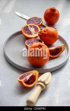 Vibrant blood oranges, both whole and sliced, arranged aesthetically on a gray concrete plate beside a wooden citrus reamer and stainless-steel knife Stock Photo