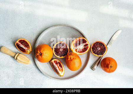 Vibrant blood oranges, both whole and sliced, arranged aesthetically on a gray concrete plate beside a wooden citrus reamer and stainless-steel knife Stock Photo