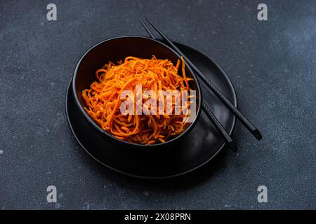 A black bowl filled with shredded carrot salad, accompanied by a pair of black chopsticks, presented on a textured dark gray surface Stock Photo