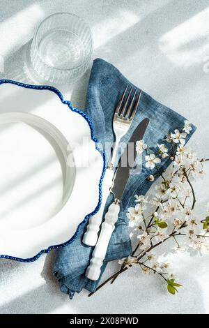 An overhead view of a stylish table setting that includes a white plate with blue rim, silver cutlery, a glass, a denim napkin, and delicate cherry bl Stock Photo