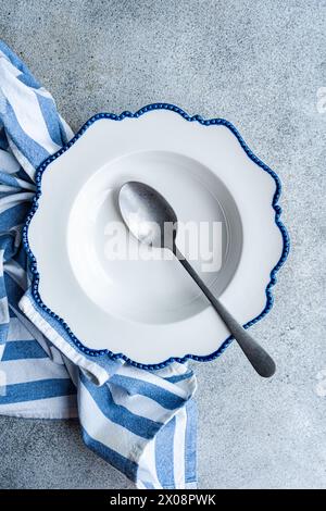 An elegant white plate with scalloped blue edges accompanied by a vintage spoon, set on a concrete background, wrapped in a striped blue tea towel Stock Photo