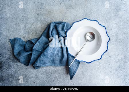 Elegant white plate with blue scalloped edges, a silver spoon, and a blue napkin set on a textured background Stock Photo