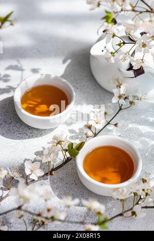 Two cups of warm tea accompanied by the delicate white flowers of cherry blossoms, casting soft shadows on a textured surface Stock Photo