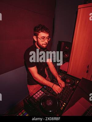 A focused DJ wearing headphones mixes tracks on a professional DJ controller, illuminated by red ambient lighting in a club-like home studio setting Stock Photo