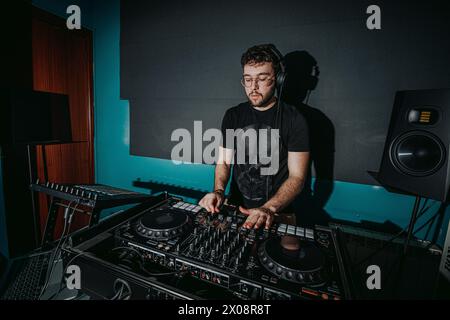 A focused male DJ mixes tracks on a professional sound mixer, with headphones and studio monitors in a dark setting Stock Photo