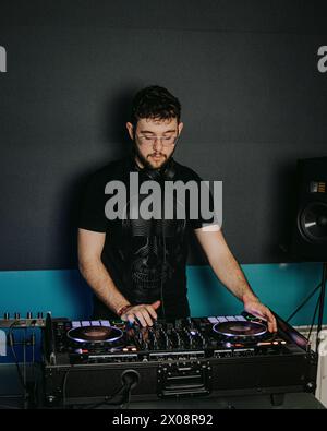 A focused young DJ with headphones around his neck mixes music on a professional DJ controller at a nightclub-like environment Stock Photo