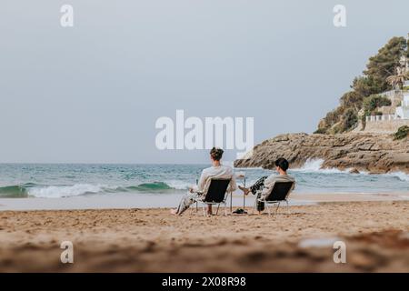 Back view of two individuals unwind in beach chairs, facing a serene sea, with waves gently lapping the shore and a rocky hill in the backdrop. Stock Photo