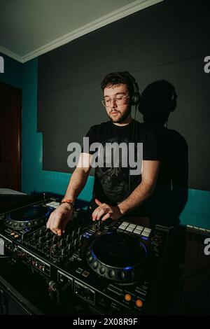 A focused DJ in a black t-shirt with a skull graphic is manipulating a sound mixer and turntable in a dimly lit room, crafting a musical experience Stock Photo