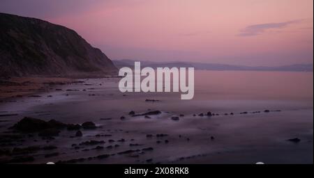 The sun's last light paints the skies and sea in shades of pink and purple at Playa de Barrika in Bilbao, with rocks ghostly in the surf Stock Photo