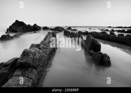 The textured rocks of Playa de Barrika stand stark against the smooth sea in this dramatic black and white coastal scene near Bilbao Stock Photo