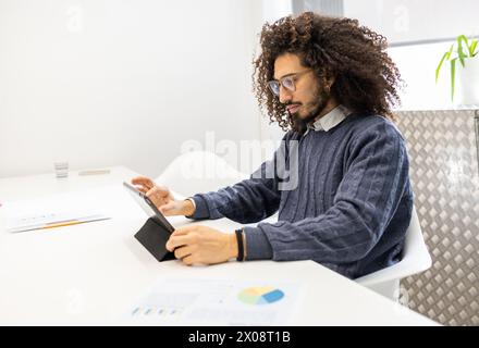 A curly-haired man with glasses, likely of mixed ethnicity, is focused on using a tablet while sitting in a white, well-lit modern office. He is weari Stock Photo