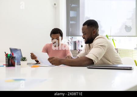 Two African American colleagues analyze documents together at a bright office workspace, with the woman reading a paper and the man focused on a digit Stock Photo