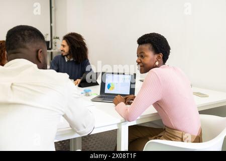 A professional African American woman in a pink sweater looks at the camera while working on a laptop in a bright office setting with colleagues engag Stock Photo