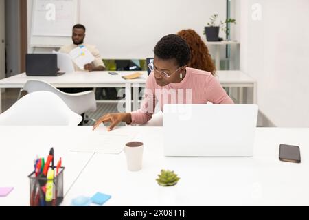 African American woman focused on paperwork in modern office, with colleagues in background. Stock Photo