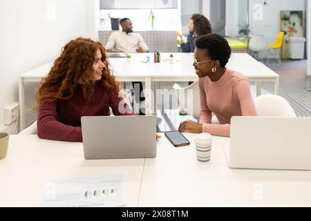 Two professional women engaging in a friendly discussion at a modern office, with colleagues in the background. Stock Photo