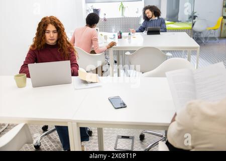 An image featuring a red-haired woman focused on her laptop, with a Black woman and a man of possible Middle Eastern descent engaged in a discussion i Stock Photo