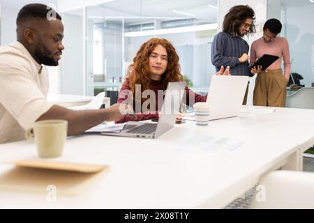 A diverse group of colleagues engages in a project discussion in a well-lit office space. A Black man and a Caucasian woman focus on a laptop, while a Stock Photo