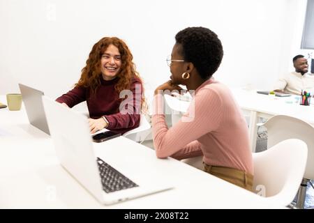 A friendly discussion between a Caucasian redhead woman and an African American woman at the office, with a man in the background. Stock Photo
