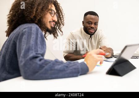 Two men, one with African ethnicity and the other with Middle Eastern or Latino ethnicity, are engaged in a discussion over a tablet in a modern offic Stock Photo