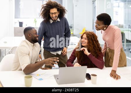 A group of four multiethnic colleagues laugh and engage in a lively discussion around a laptop in an office setting. Stock Photo