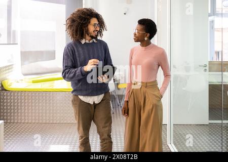 Two multiethnic colleagues, a Black woman and a man, are engaged in a friendly discussion in a brightly lit office. The man is holding a tablet and bo Stock Photo