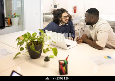 Two men in a bright office, one with Middle Eastern traits and the other Black, discussing work and looking at each other with a laptop and plants on Stock Photo
