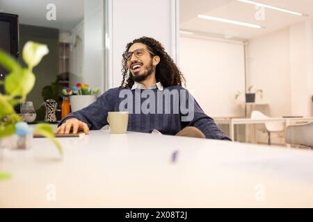 Cheerful man with curly hair smiling and looking away in an office environment, holding a coffee mug. Stock Photo