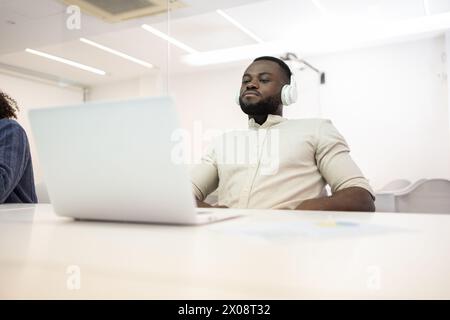 African American man working on his laptop at a bright office, wearing headphones and looking away from the camera. Stock Photo