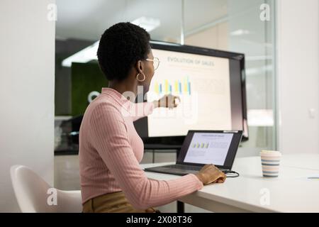 African American woman analyzing sales data on a computer monitor with a laptop and coffee cup on her desk. Stock Photo