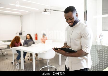 Cheerful African American man looking at digital tablet with coworkers in blurred background. Stock Photo