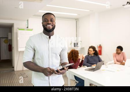 Smiling African American man looking at camera in office, coworkers collaborating in the background. Stock Photo