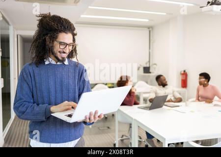 A cheerful young man with Afro-textured hair is engrossed in his laptop, standing in a modern office setting. His colleagues, diverse in ethnicity, en Stock Photo