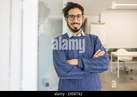 Confident Middle Eastern man with a warm smile, arms crossed, looking at the camera in a bright office setting. Stock Photo