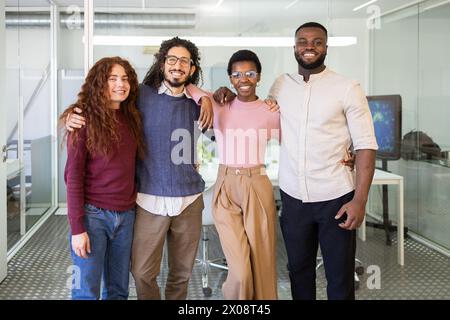 Four diverse colleagues sharing a friendly moment, standing with arms around each other in a modern office setting, looking at the camera and smiling. Stock Photo
