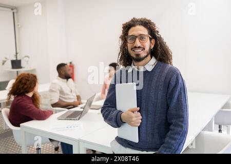 A Middle Eastern man smiling at the camera, holding a tablet, with coworkers in a modern office setting behind him. Stock Photo