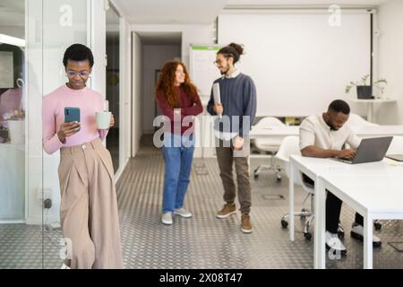 Diverse office scene with an African American woman using her phone and holding a mug, coworkers chatting, and a man on a laptop. Stock Photo