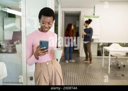 A young Black woman is engaging with her smartphone while holding a coffee mug in a contemporary office space, with coworkers in the background. Stock Photo
