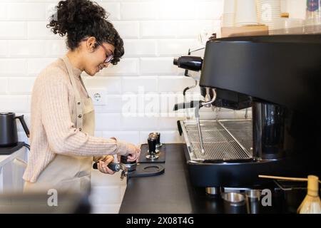 A professional hispanic woman barista tamps ground coffee into a portafilter at an espresso machine in a coffee setting, showcasing the coffee-making Stock Photo