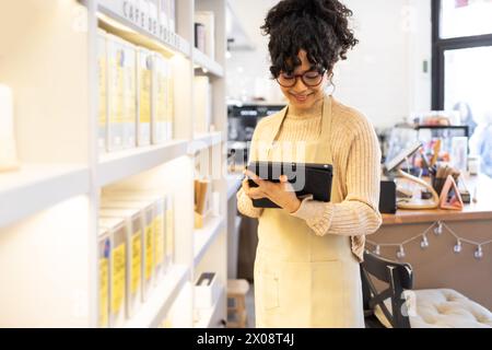 A cheerful small business owner stands in her boutique, browsing on a digital tablet amidst shelves stocked with products. Stock Photo