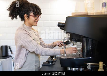 A smiling hispanic female barista expertly operates a professional espresso machine in a bright and modern cafe setting. Stock Photo