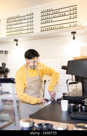 A focused barista in a yellow sweater and apron works on an espresso machine in a well-lit coffee with a menu board in the background. Stock Photo