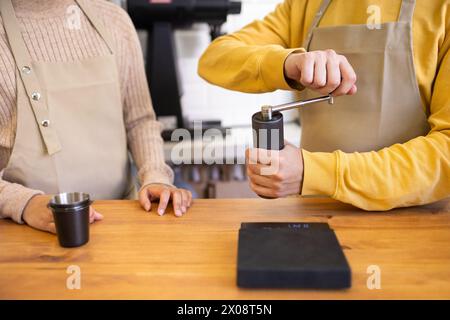 Close-up of a barista in an apron using a hand-operated coffee grinder at a wooden counter. Stock Photo