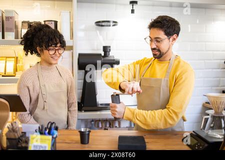 Two cheerful multiethnic baristas in aprons are happily engaging in their duties at a modern, well-lit coffee house, showcasing teamwork and customer Stock Photo