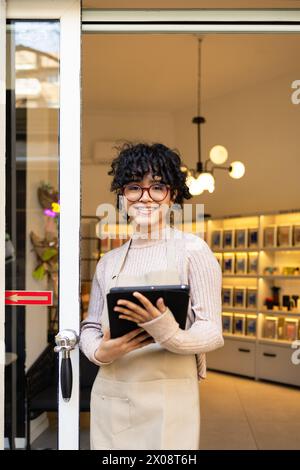 A cheerful entrepreneur stands at the entrance of her boutique, holding a digital tablet, ready to welcome customers. Stock Photo