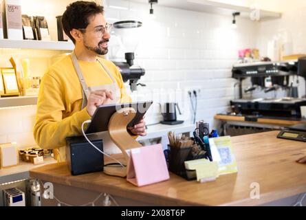 A cheerful male coffee owner operates a digital tablet behind the counter with coffee equipment in the background. Stock Photo