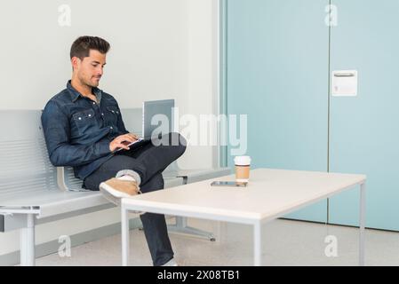 Young man using laptop and having coffee sitting on chair in hall. Stock Photo