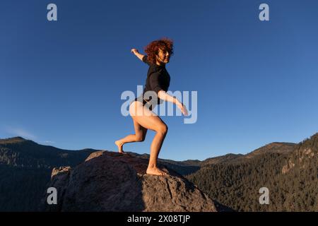A dynamic image capturing a contemporary dancer striking a poised pose on a rock against a breathtaking mountainous backdrop under a clear blue sky Stock Photo