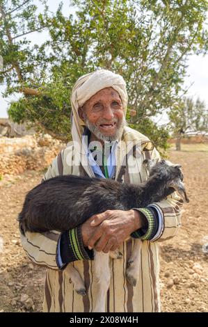 A senior Moroccan man in traditional attire smiling while cradling a black goat in a rural landscape, showcasing local lifestyle and culture Stock Photo